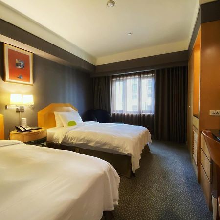 Ambassador Hotel Καοσιούνγκ Εξωτερικό φωτογραφία The photo shows a hotel room with two beds, each made up with white linens. The walls are painted in dark colors, while the bedding and furniture are lighter in tone. There is a nightstand between the beds with a lamp and telephone on it. A window wi
