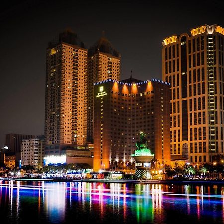 Ambassador Hotel Καοσιούνγκ Εξωτερικό φωτογραφία The photo depicts a vibrant nighttime cityscape featuring several tall buildings illuminated by colorful lights. Prominent structures include a large hotel and other high-rise buildings with illuminated facades. In front of these buildings, there app