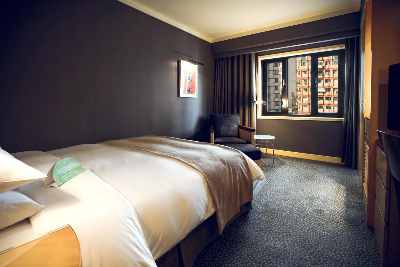 Ambassador Hotel Καοσιούνγκ Εξωτερικό φωτογραφία The photo shows a hotel room featuring a large bed with a light-colored duvet. The walls are painted in a dark hue, creating a cozy atmosphere. There is a chair and a small side table near the window, which is covered by curtains. Through the window,