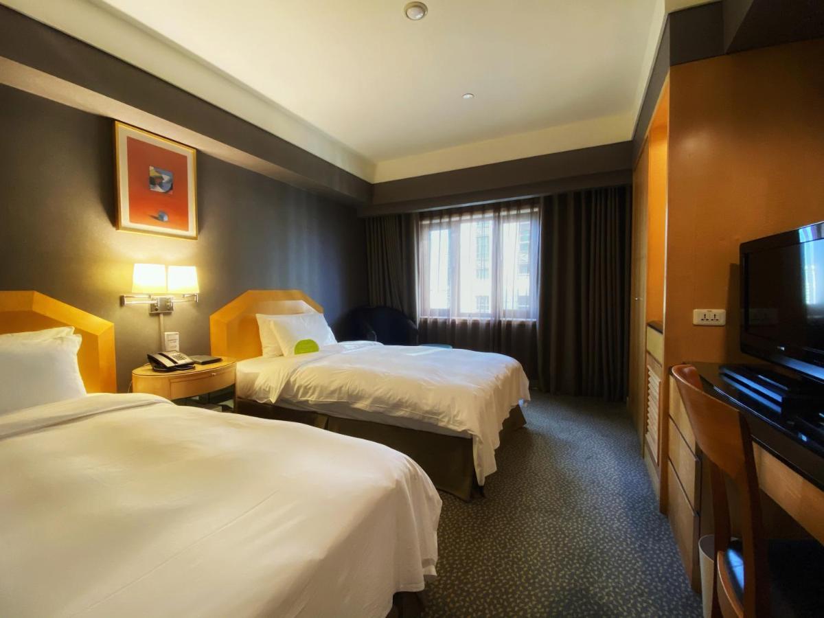 Ambassador Hotel Καοσιούνγκ Εξωτερικό φωτογραφία The photo shows a hotel room with two beds, each made up with white linens. The walls are painted in dark colors, while the bedding and furniture are lighter in tone. There is a nightstand between the beds with a lamp and telephone on it. A window wi