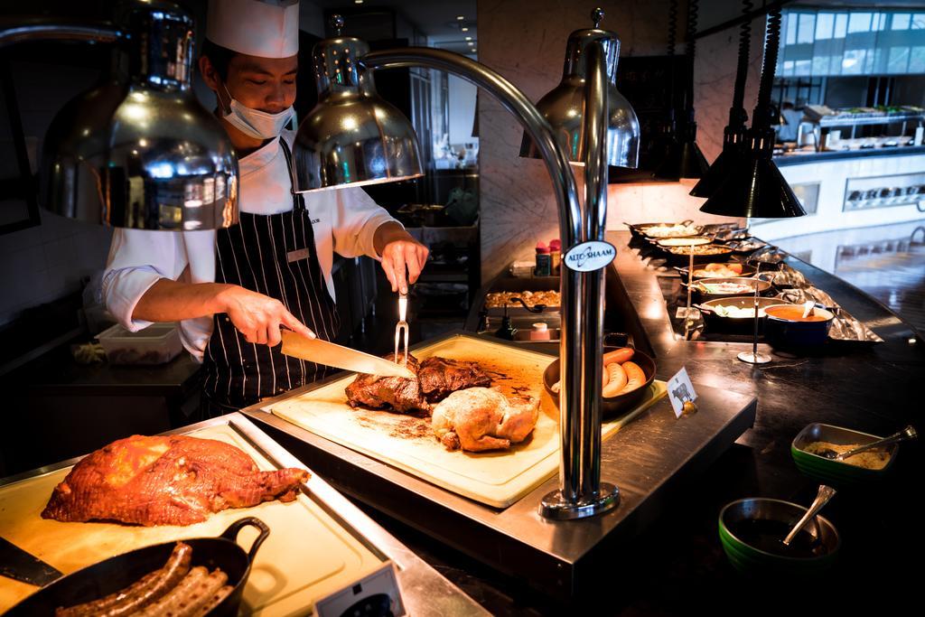 Ambassador Hotel Καοσιούνγκ Εξωτερικό φωτογραφία The photo depicts a chef in a kitchen or buffet setting. The chef, wearing a white chef's hat and a striped apron, is slicing meat on a cutting board. There are various meat dishes displayed under warming lamps, indicating a buffet-style layout. You 