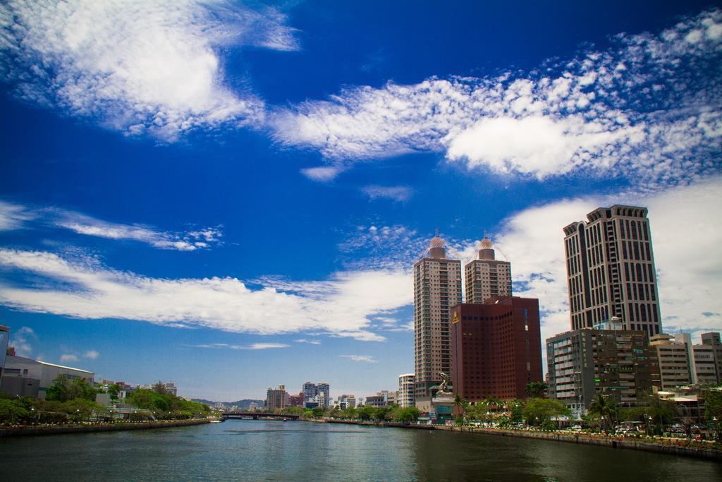 Ambassador Hotel Καοσιούνγκ Εξωτερικό φωτογραφία The photo showcases a scenic view of a river flanked by modern skyscrapers. The buildings have a mix of architectural styles, including tall, glass-fronted structures and a reddish-brown building. In the background, a clear blue sky features fluffy w