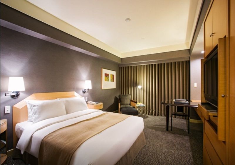 Ambassador Hotel Καοσιούνγκ Εξωτερικό φωτογραφία The photo shows a modern hotel room featuring a neatly made bed with white linens and a beige bedspread. There are bedside lamps on either side of the bed. One corner of the room includes a comfortable armchair, and adjacent to it is a small table. T