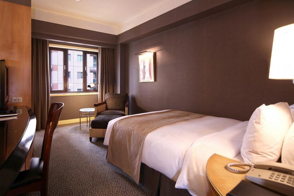 Ambassador Hotel Καοσιούνγκ Εξωτερικό φωτογραφία The photo shows a hotel room featuring a neatly made single bed with a light-colored blanket. There is a comfortable chair in the corner alongside a small round table. A large window allows natural light to enter the room, and the walls are painted i