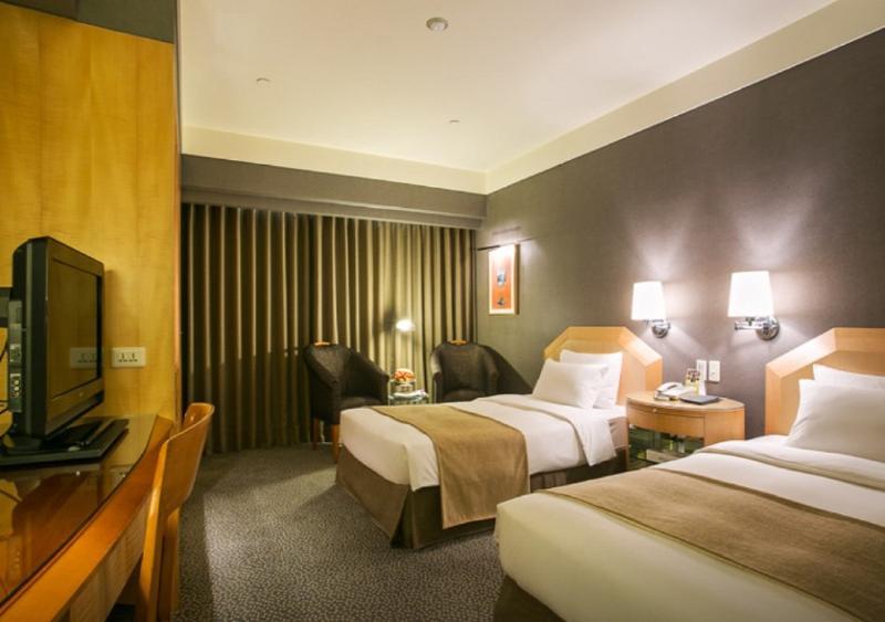 Ambassador Hotel Καοσιούνγκ Εξωτερικό φωτογραφία The photo shows a hotel room featuring two single beds with light-colored bedding. The walls are painted in a muted color, and there are two wall-mounted lamps above each bed. A wooden desk and chair are positioned against one wall, accompanied by a 