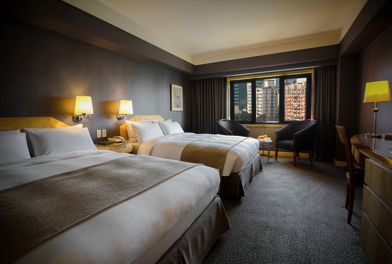 Ambassador Hotel Καοσιούνγκ Εξωτερικό φωτογραφία The photograph shows a hotel room featuring two double beds with white linens and light brown accents. The walls are painted a dark color, and there are bedside lamps beside each bed. A window offers a view of the outside, and there are two chairs po