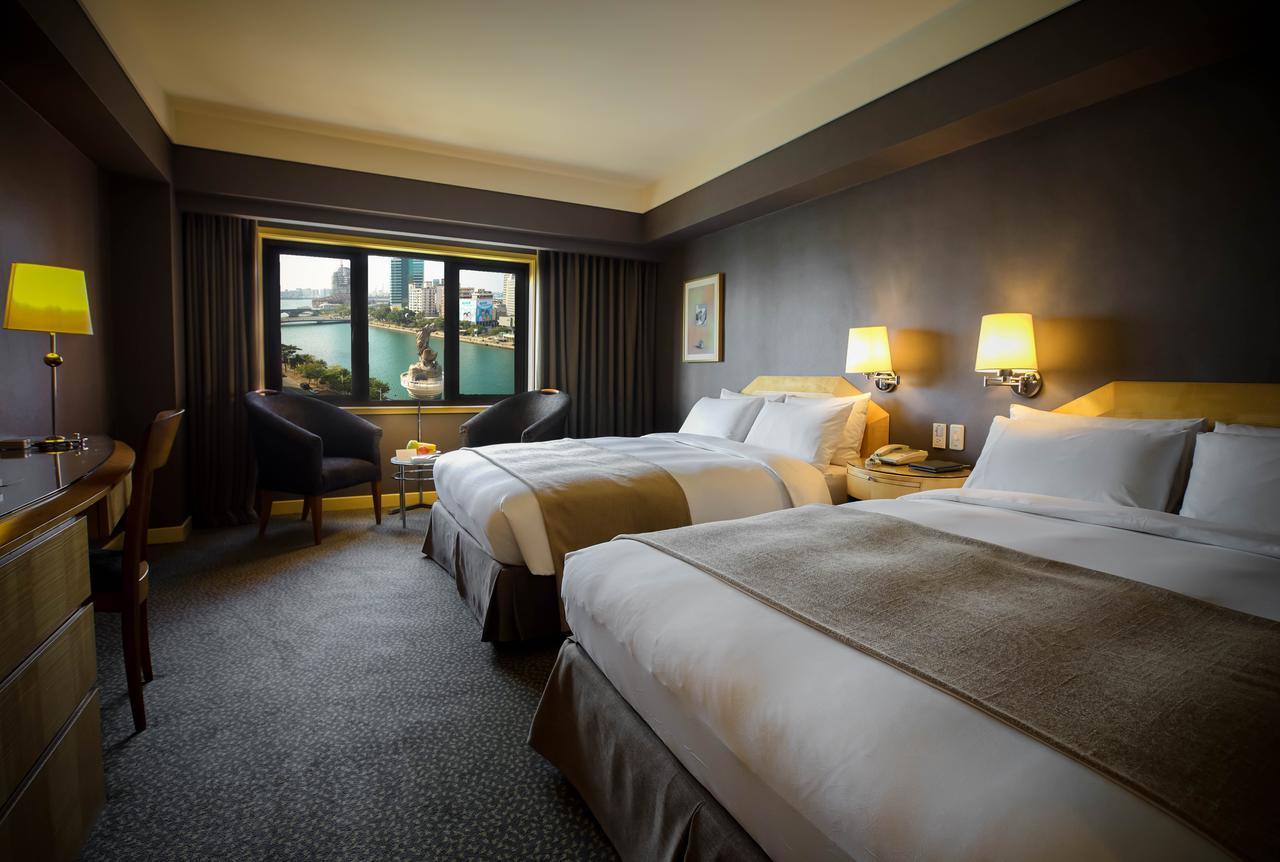 Ambassador Hotel Καοσιούνγκ Εξωτερικό φωτογραφία The photo shows a hotel room featuring two large beds with white linens and beige accents. The room has a modern design with dark walls and plush carpeting. There are lamps on each side of the beds, providing warm lighting. A large window offers a vi