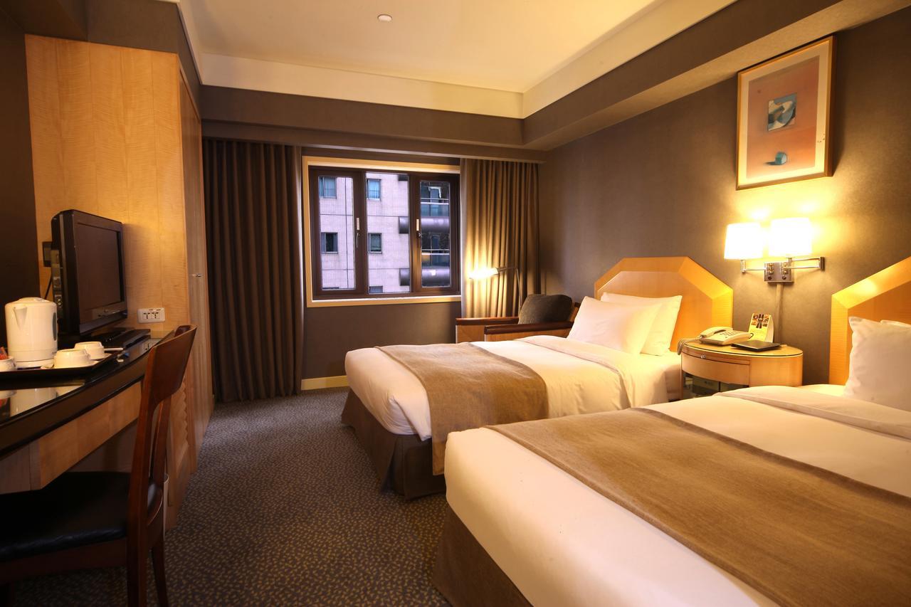 Ambassador Hotel Καοσιούνγκ Εξωτερικό φωτογραφία The photo shows a hotel room featuring two twin beds with neutral-colored bed linens. The room has a modern design, with warm lighting and wooden furnishings. There's a desk and chair positioned against one wall, along with a television. A large wind
