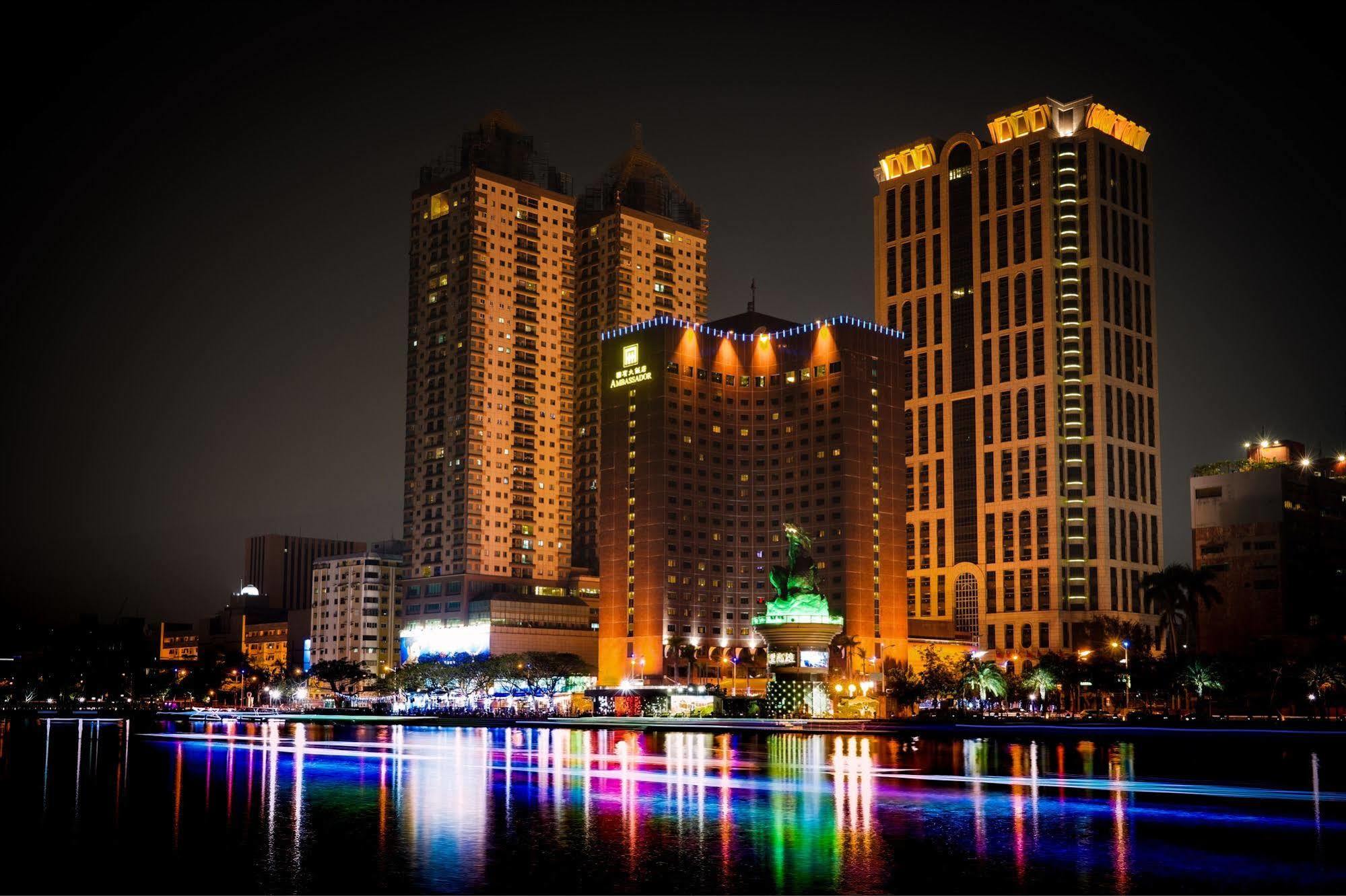 Ambassador Hotel Καοσιούνγκ Εξωτερικό φωτογραφία The photo depicts a vibrant nighttime cityscape featuring several tall buildings illuminated by colorful lights. Prominent structures include a large hotel and other high-rise buildings with illuminated facades. In front of these buildings, there app