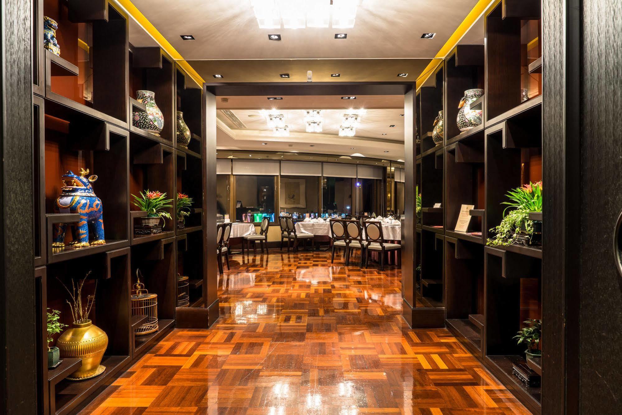 Ambassador Hotel Καοσιούνγκ Εξωτερικό φωτογραφία The photo shows the interior of a restaurant or dining area. It features a corridor lined with dark wooden shelves displaying decorative items and plants. The flooring is made of polished wood, which reflects the warm lighting from the ceiling fixtur