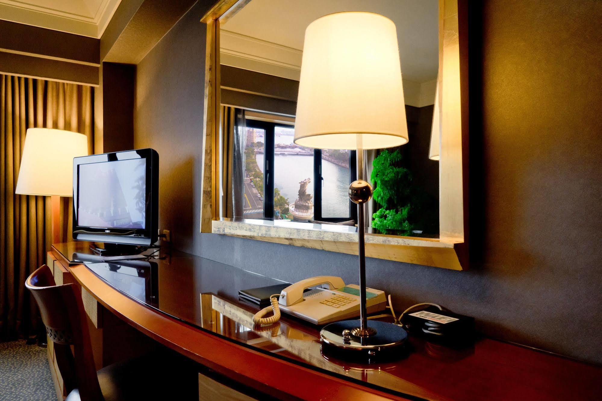 Ambassador Hotel Καοσιούνγκ Εξωτερικό φωτογραφία The photo depicts a hotel room interior featuring a sleek wooden desk with a curved design. On the desk, there is a modern lamp with a white shade, a telephone, and a television set to the left. A mirror hangs above the desk, reflecting light and the
