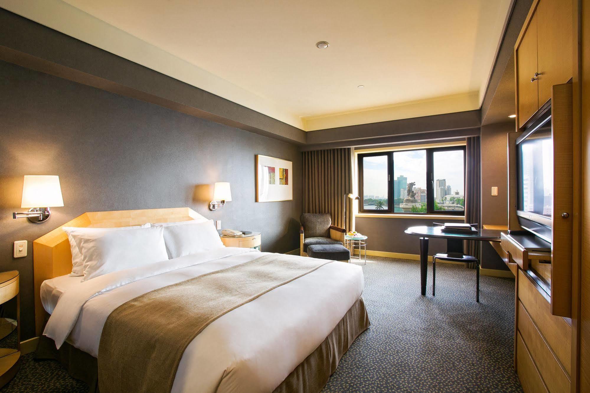 Ambassador Hotel Καοσιούνγκ Εξωτερικό φωτογραφία The photo shows a hotel room that features a large bed with crisp white linens and a beige throw blanket. There are bedside lamps on either side of the bed. A comfortable chair is positioned near a window with light drapes, and a small desk with a ch