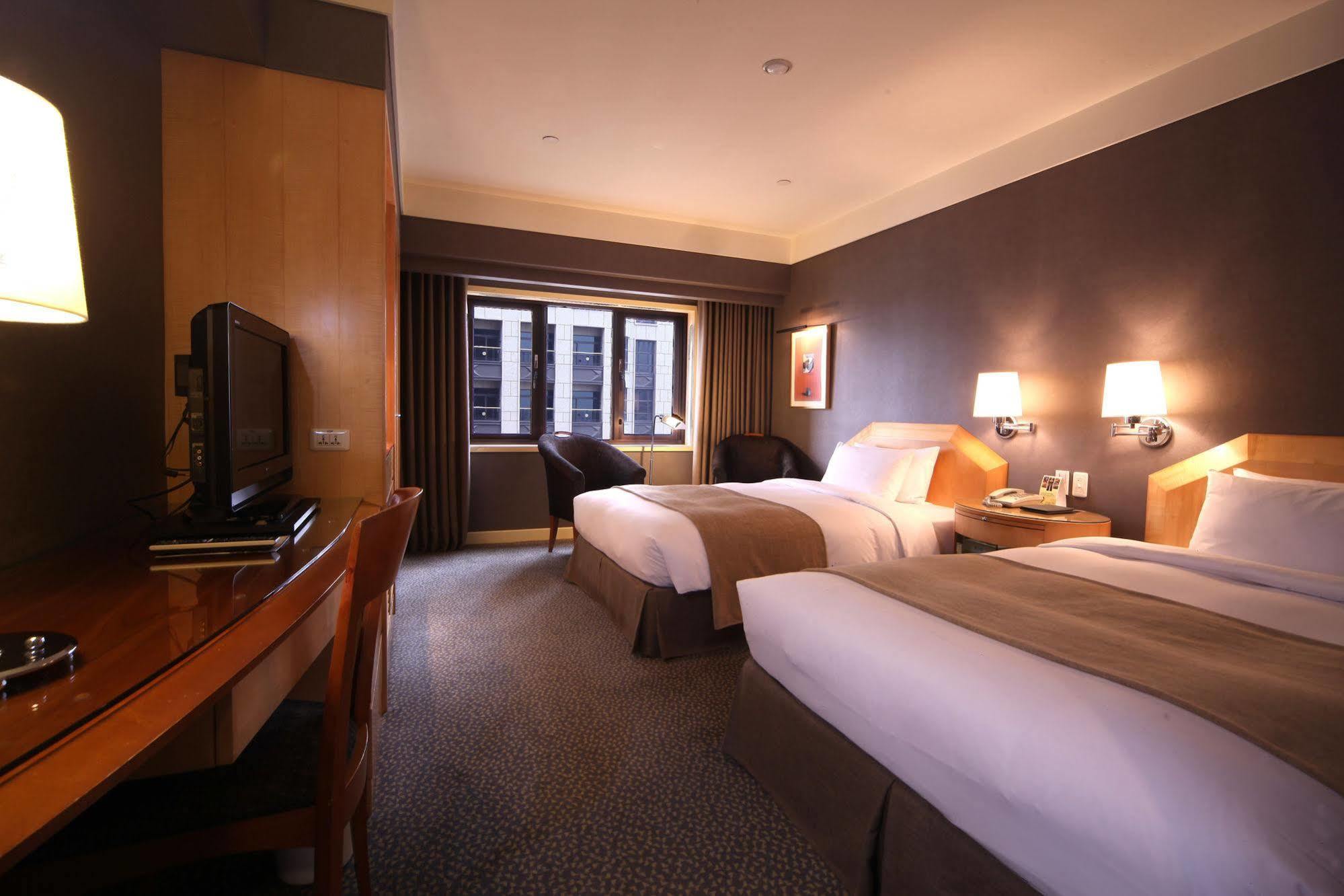 Ambassador Hotel Καοσιούνγκ Εξωτερικό φωτογραφία The photo shows a hotel room featuring two twin beds, each with light-colored bedding. There are bedside lamps on each side of the beds. A wooden desk with a chair is positioned next to a wall-mounted television. The room has a large window with shee