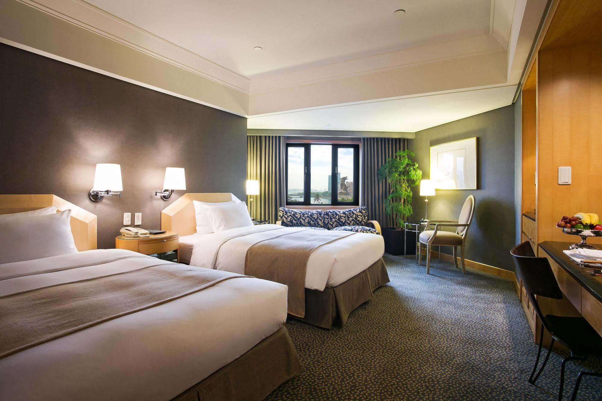 Ambassador Hotel Καοσιούνγκ Εξωτερικό φωτογραφία The photo shows a hotel room featuring two neatly made beds with white bedding and light brown accents. There are bedside lamps on either side of the beds. A large window lets in natural light, and there’s a small sitting area with a couch and a plan