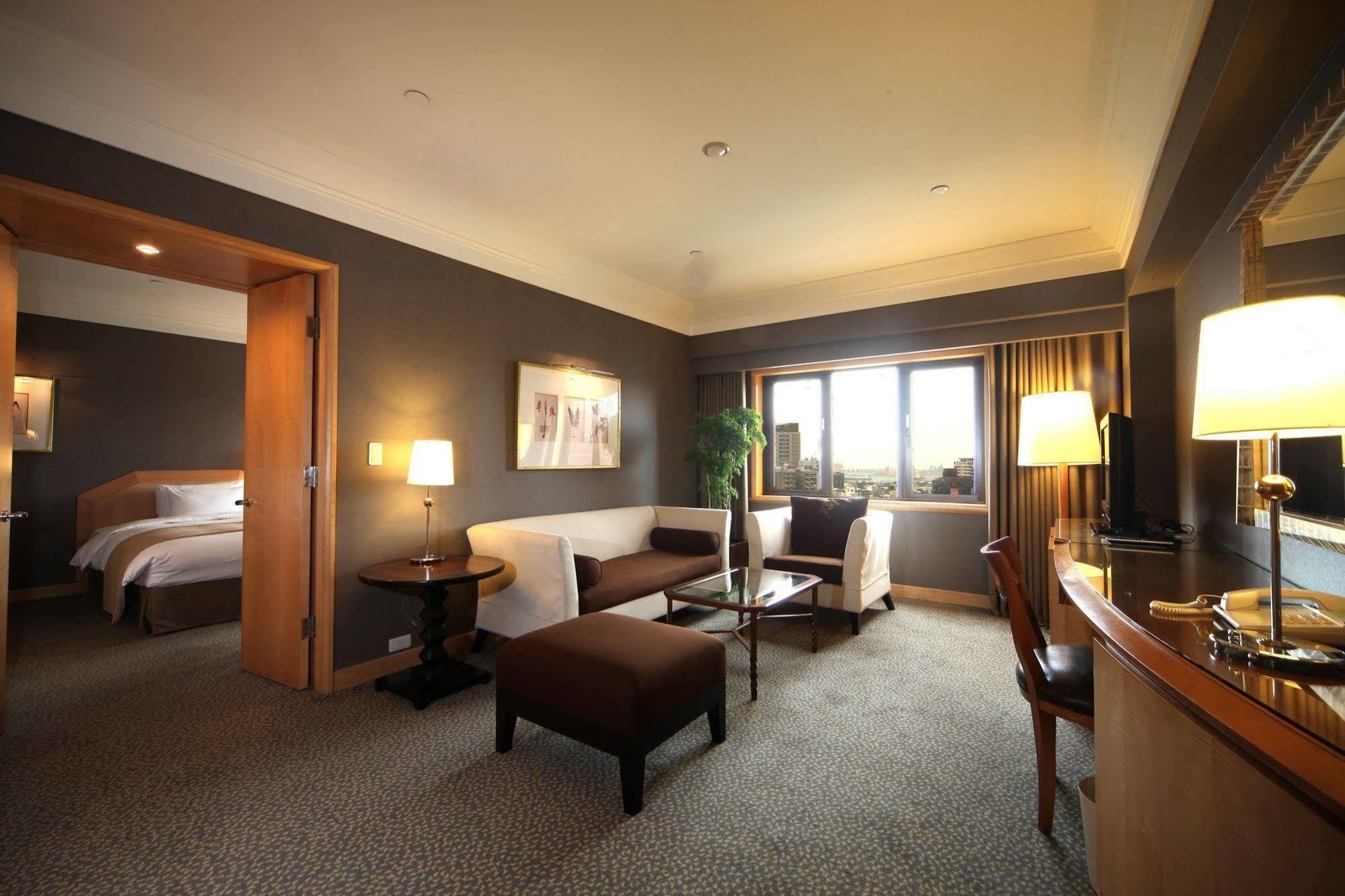 Ambassador Hotel Καοσιούνγκ Εξωτερικό φωτογραφία The photo shows an elegantly furnished hotel room. The space features a comfortable seating area with a white sofa and a dark brown ottoman placed in front of a glass coffee table. There is a small side table with a lamp beside the sofa. A large wind