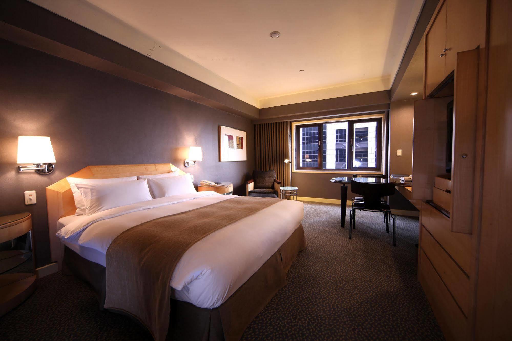 Ambassador Hotel Καοσιούνγκ Εξωτερικό φωτογραφία The photo shows a hotel room featuring a large bed with a white bedspread and a brown accent. There are nightstands on either side of the bed, each with a lamp. A chair is positioned near a window, which offers a view outside. A wooden desk and chair