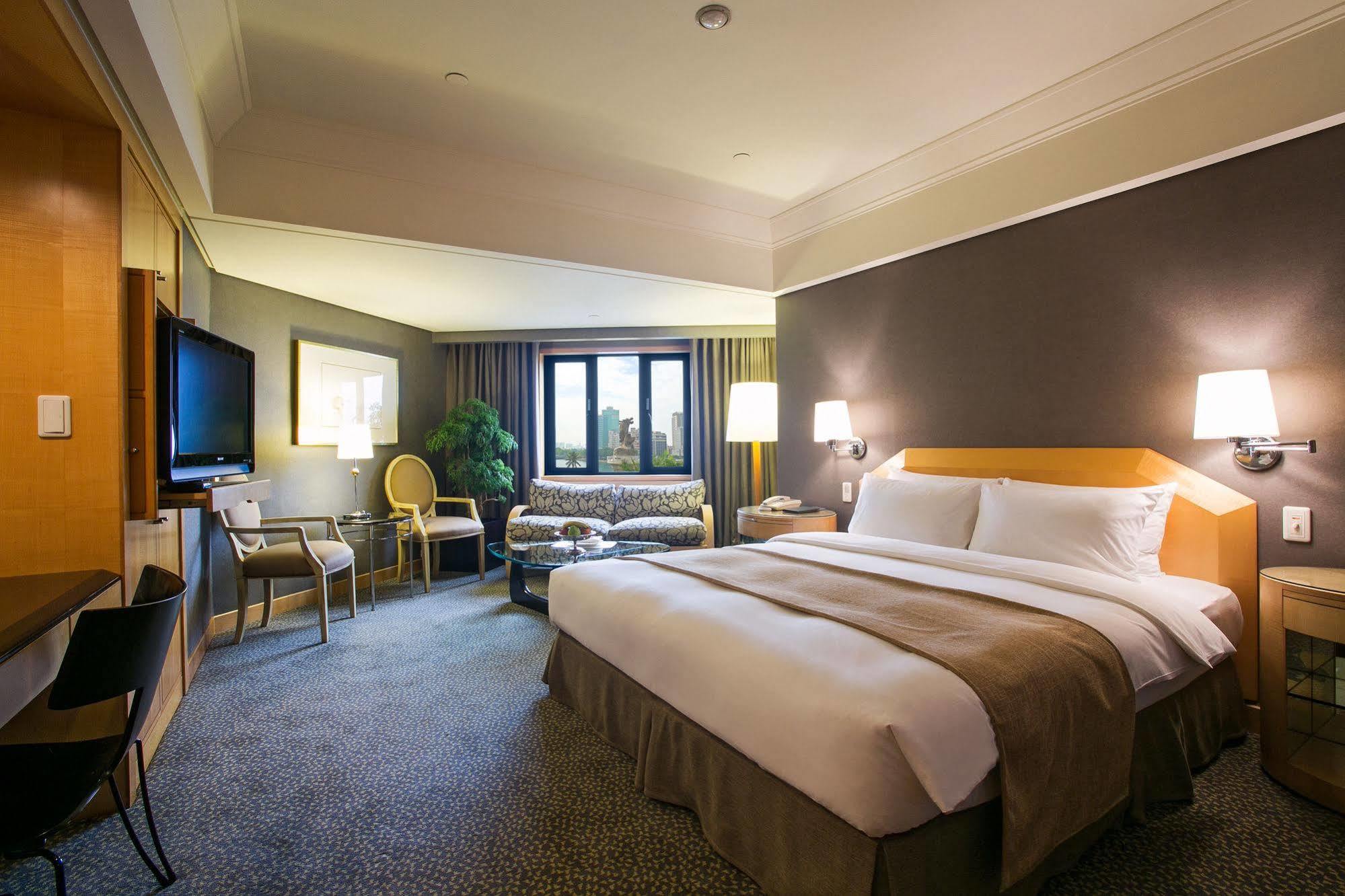 Ambassador Hotel Καοσιούνγκ Εξωτερικό φωτογραφία The photo shows a well-appointed hotel room. It features a large bed with white and beige bedding, a comfortable seating area with a sofa and chairs, and a small coffee table. A flat-screen television is mounted on the wall opposite the bed. There ar