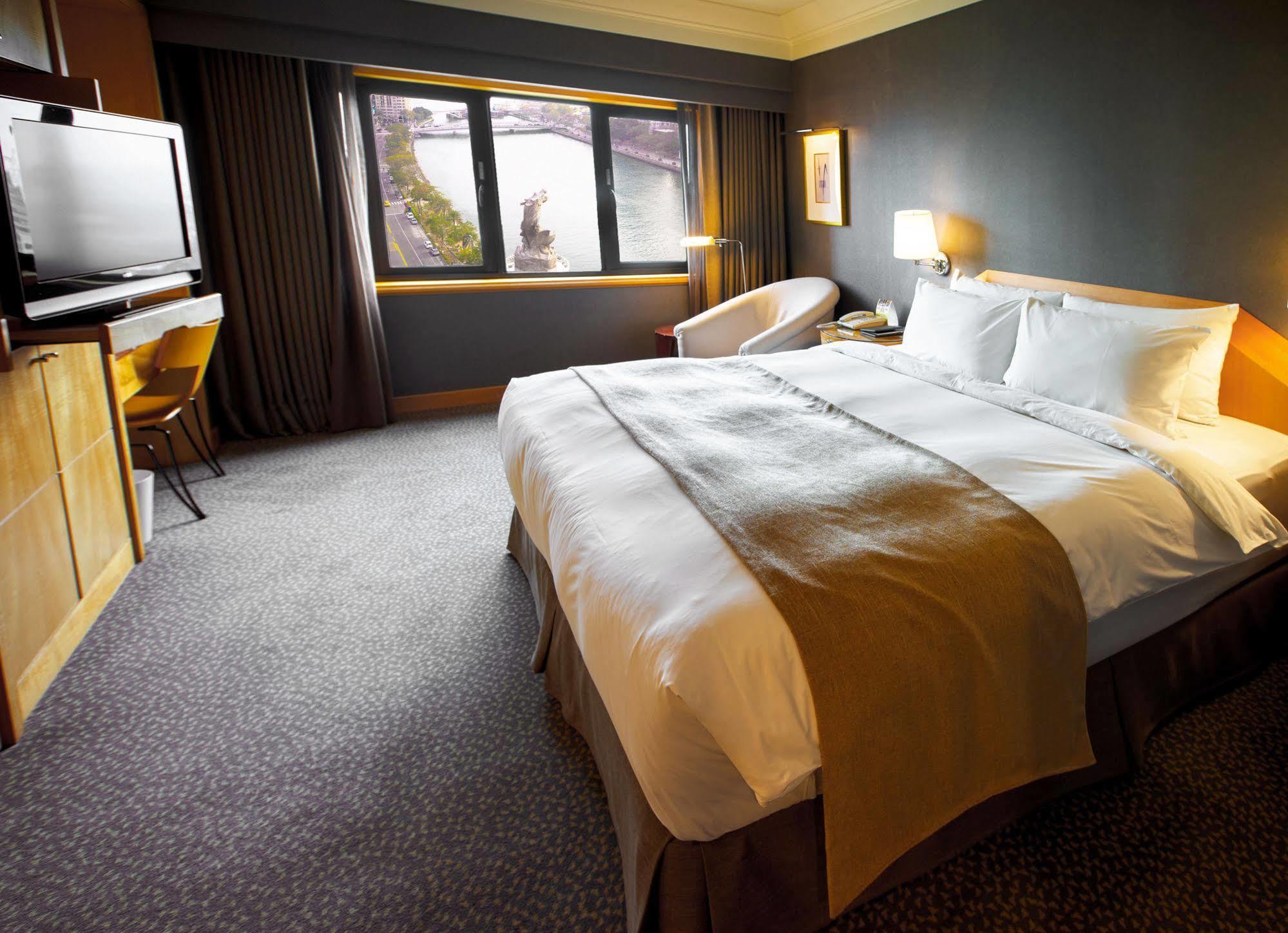 Ambassador Hotel Καοσιούνγκ Εξωτερικό φωτογραφία The photo depicts a modern hotel room featuring a spacious layout. In the center, there is a large bed with crisp white linens and a decorative brown throw blanket. To the side, there is a comfortable armchair and a small table. A television is mount