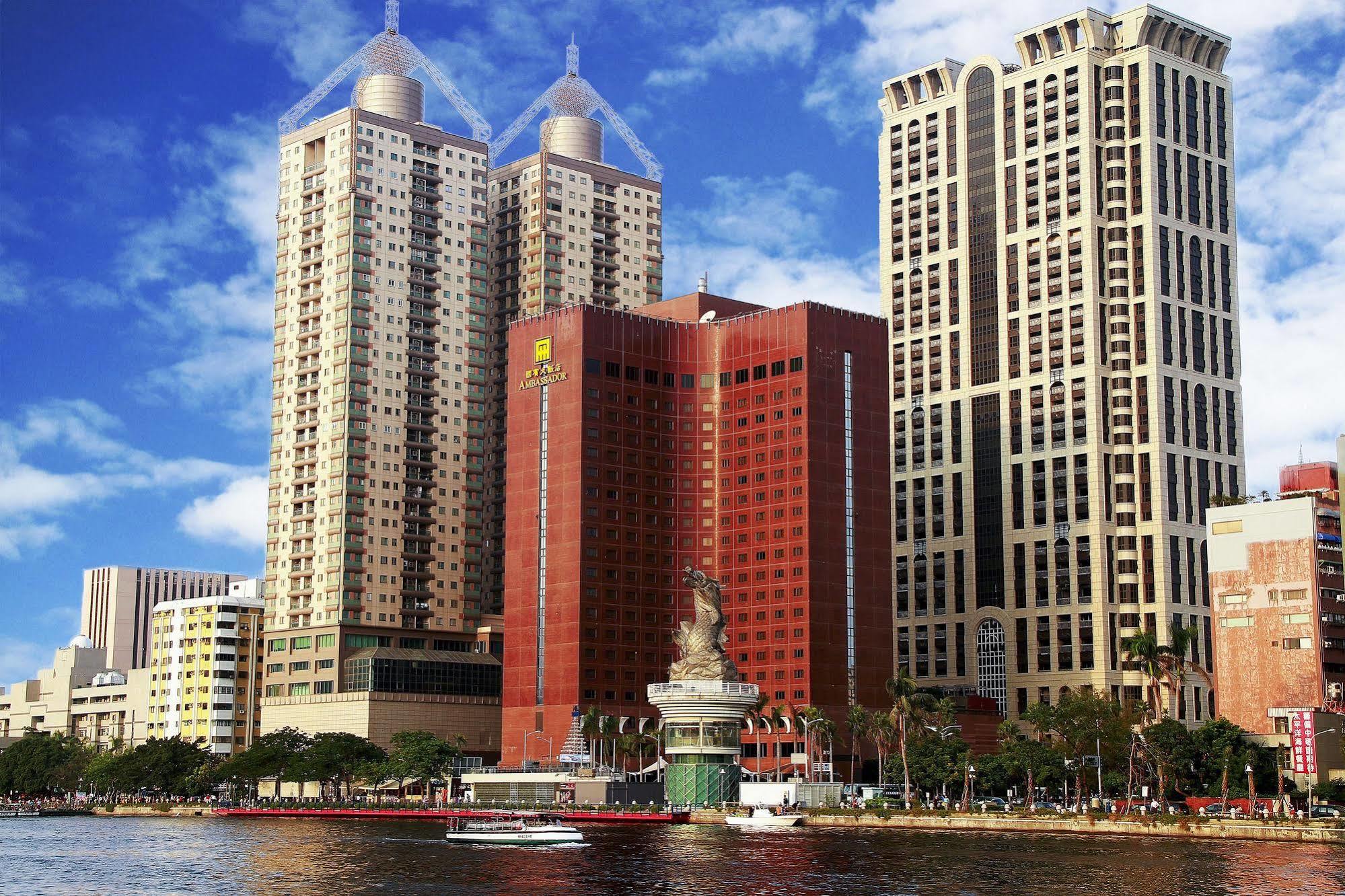 Ambassador Hotel Καοσιούνγκ Εξωτερικό φωτογραφία The photo shows a cityscape featuring several tall buildings, including two prominent skyscrapers with distinctive architectural features. The structures are situated near a body of water, suggesting a waterfront location. The sky is blue with some c