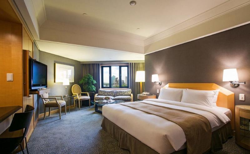 Ambassador Hotel Καοσιούνγκ Εξωτερικό φωτογραφία The photo shows a hotel room that features a large bed with white linens and a light brown throw blanket. There are two bedside lamps on either side of the bed. In the corner, there is a cozy seating area with a patterned sofa and two chairs. A telev
