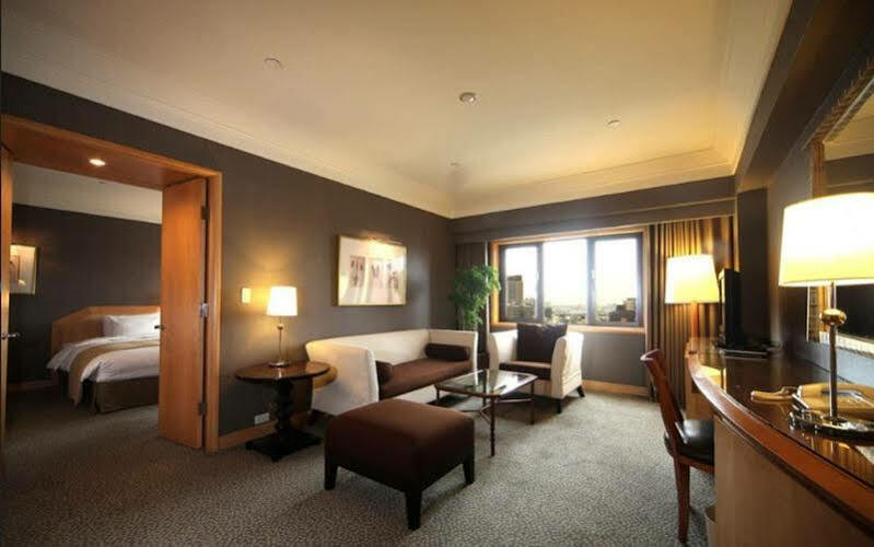 Ambassador Hotel Καοσιούνγκ Εξωτερικό φωτογραφία The photo shows a stylish hotel room with modern décor. There is a comfortable seating area featuring a white sofa and brown ottoman, along with a glass coffee table. A side table with a lamp is positioned next to the sofa. The room has a large windo
