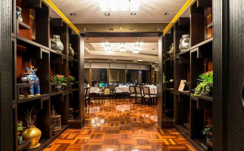Ambassador Hotel Καοσιούνγκ Εξωτερικό φωτογραφία The photo shows an elegant restaurant interior. In the foreground, there are dark wooden shelves filled with decorative items, such as vases and small sculptures, along with plants. The floor is made of polished wood, featuring a geometric pattern. I